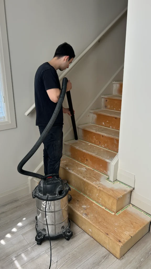 An installer using a heavy-duty vacuum to clean debris and dust from bare wooden stairs after removing old carpet, ensuring a smooth surface for the new underlay.